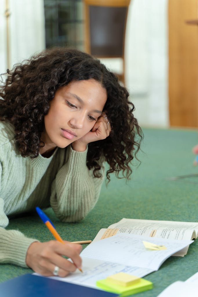 Black woman in a cozy sweater studying on the floor, surrounded by textbooks and notes, deep in thought.